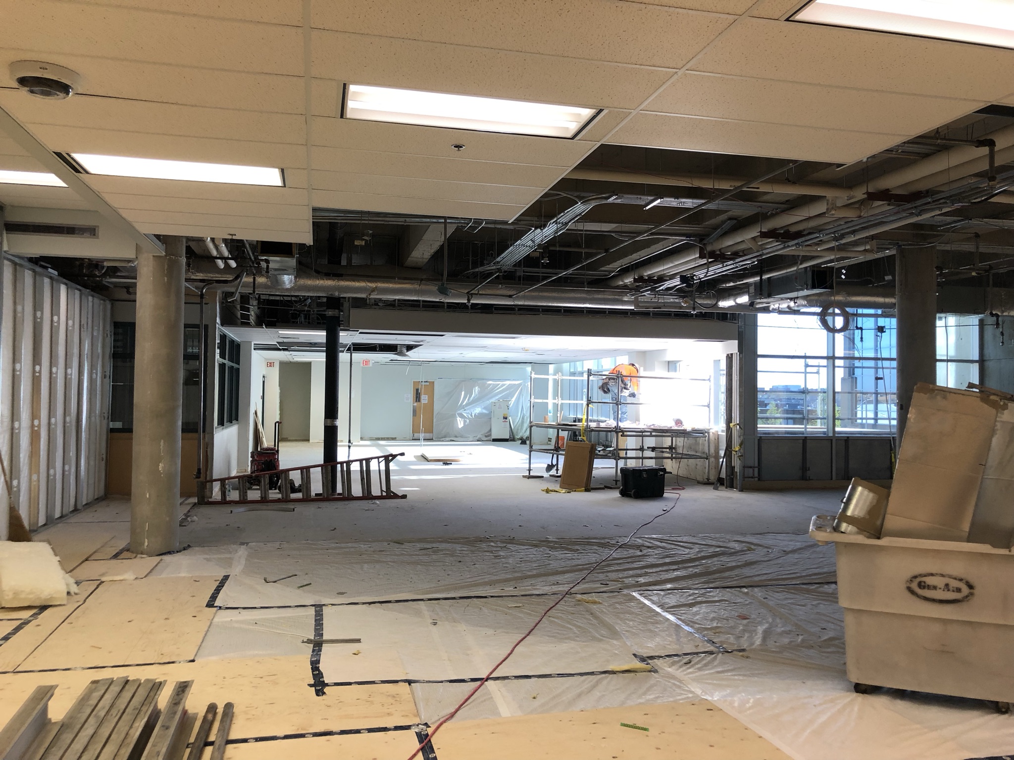 A photograph of the space where the library service desk was. It is now demolished and the wall behind it has been removed to revel the space behind it. Flooring and ceiling have been removed and expose the concrete and wiring underneath.