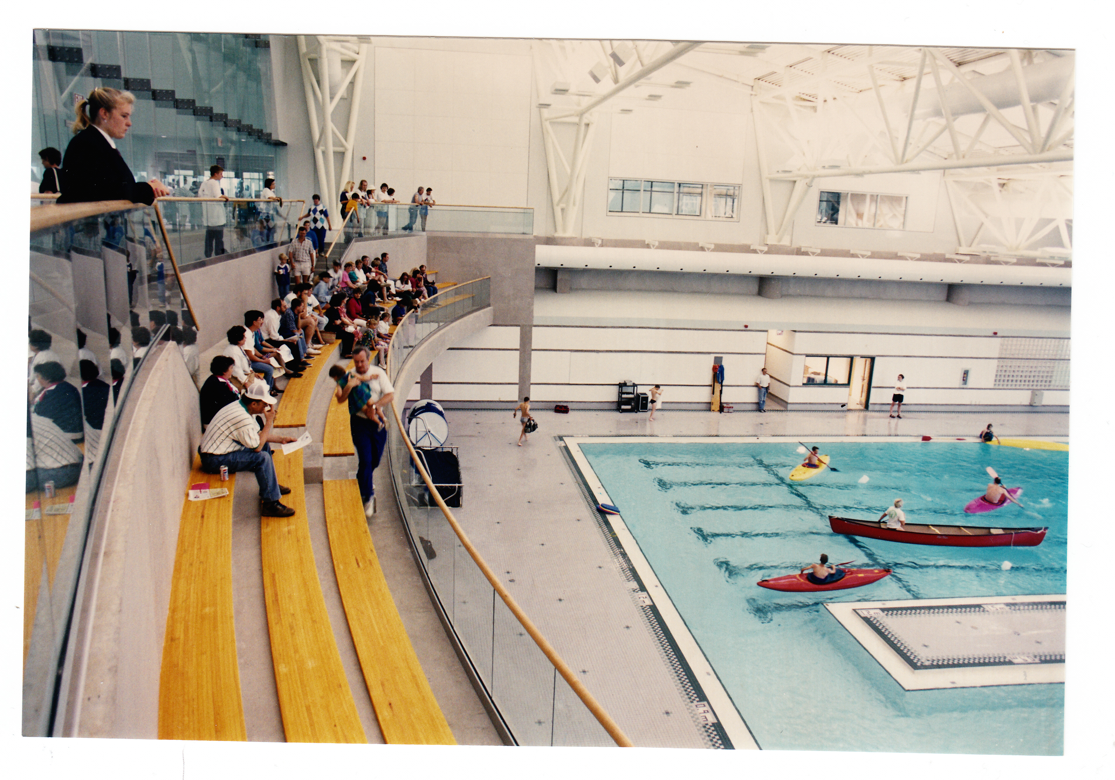 An old photograph of the opening day of the Sport and Wellness Centre. The photo is taken from the bleachers overlooking the pool. Many people are sitting in the bleachers while a few people are paddling in kayaks and canoes in the pool.
