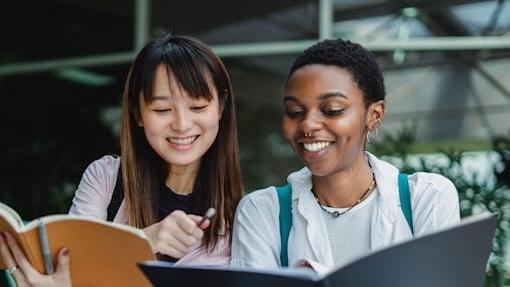 Two students sitting beside each other smile at their open notebooks.